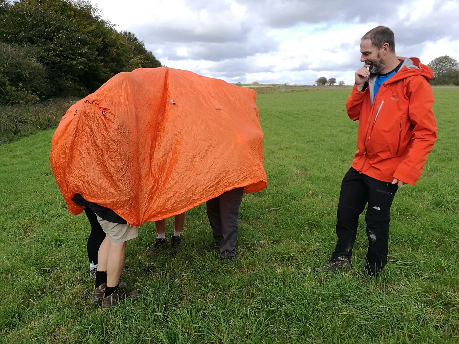 Team members sheltering together beneath an orange survival bag